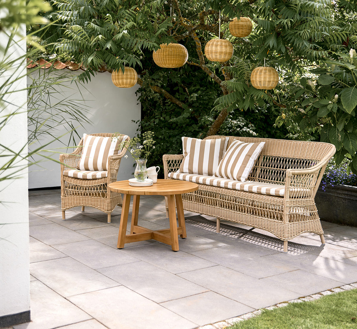 Dies ist ein Lifestyle-Bild von Charlot 3-Sitzer-Sofa in natural ArtFibre Aluminium, auf einer Küstenterrasse mit Schilfdach und Meerblick.