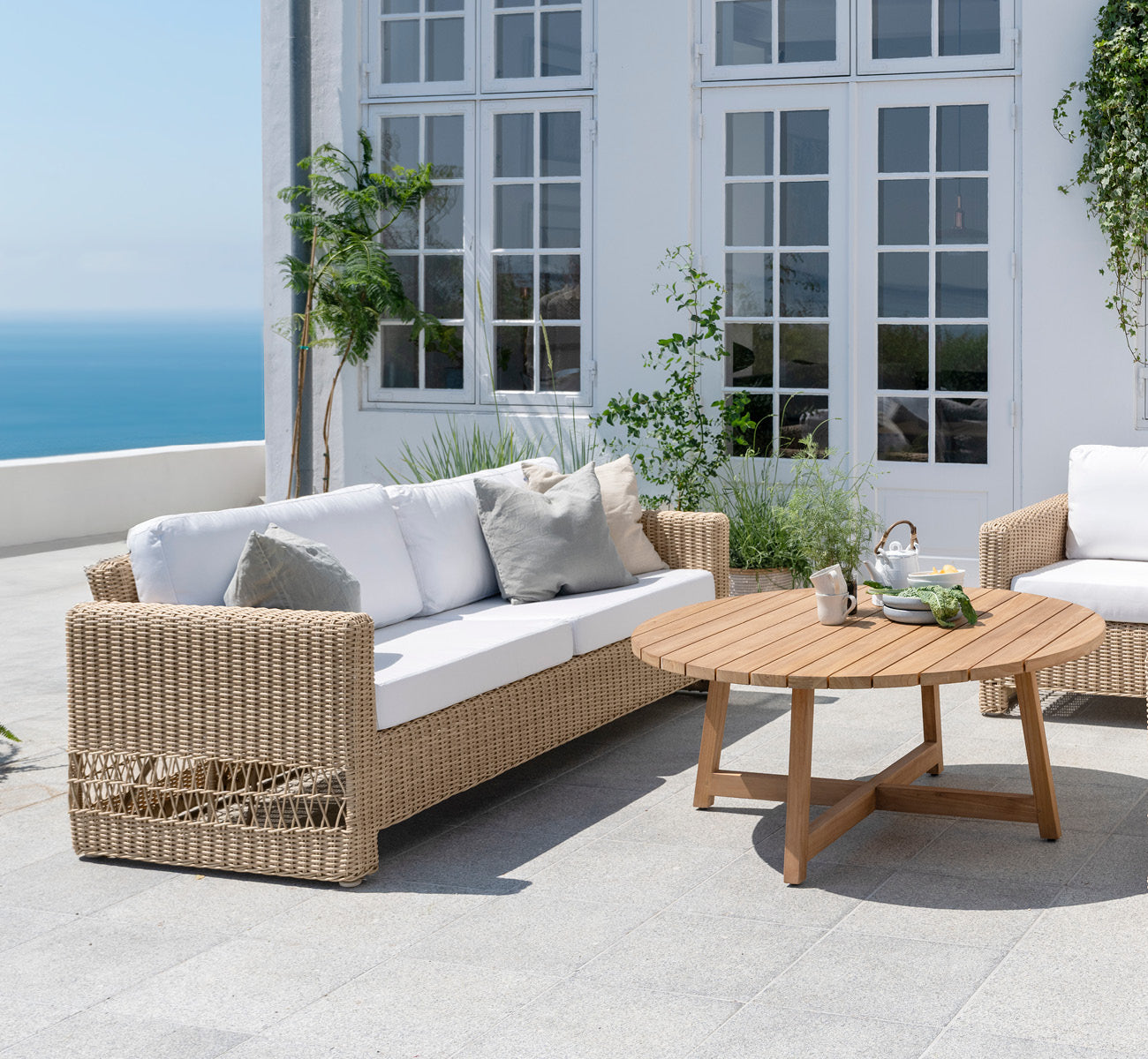Dies ist ein Lifestyle-Bild von Carrie Sofa in natural ArtFibre Aluminium, auf einer Küstenterrasse mit weißem Holzdeck, Meerblick und Pergola.