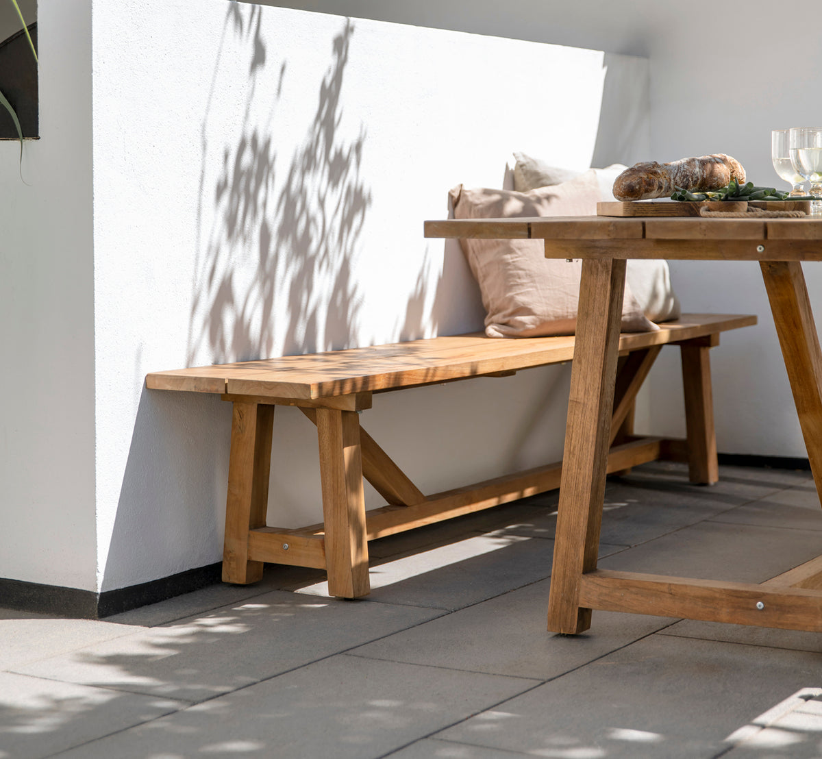 Dies ist ein Lifestyle-Bild von George Außenbank in outdoor Teak, auf einer Außenterrasse mit grauem Fliesenboden und einer weiß verputzten Mauer.