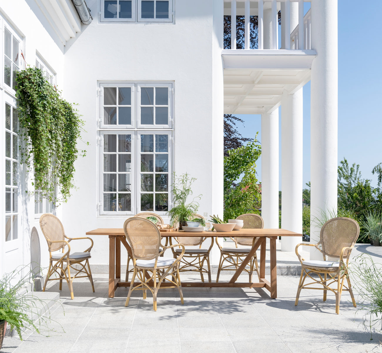 Dies ist ein Lifestyle-Bild von George Außentisch 240 in outdoor Teak, auf einer Gartenterrasse im Freien mit weißer Hausfassade und Bepflanzung.