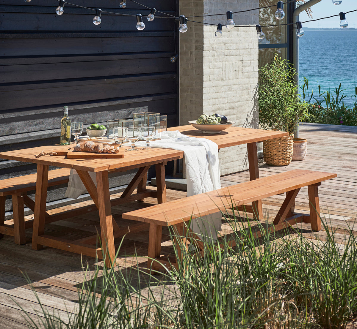 Dies ist ein Lifestyle-Bild von George Außentisch 240 in outdoor Teak, auf einer Gartenterrasse im Freien mit weißer Hausfassade und Bepflanzung.