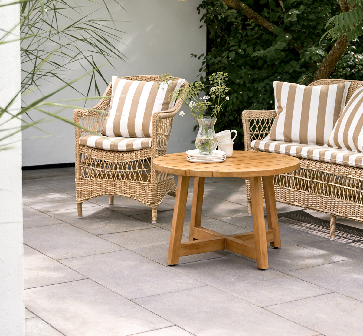 Dies ist ein Lifestyle-Bild von Victor Teak Lounge Table Ø75 cm in teak outdoor. Auf einer Strandterrasse zwischen Tagesbetten mit gestreiften Kissen aufgestellt.
