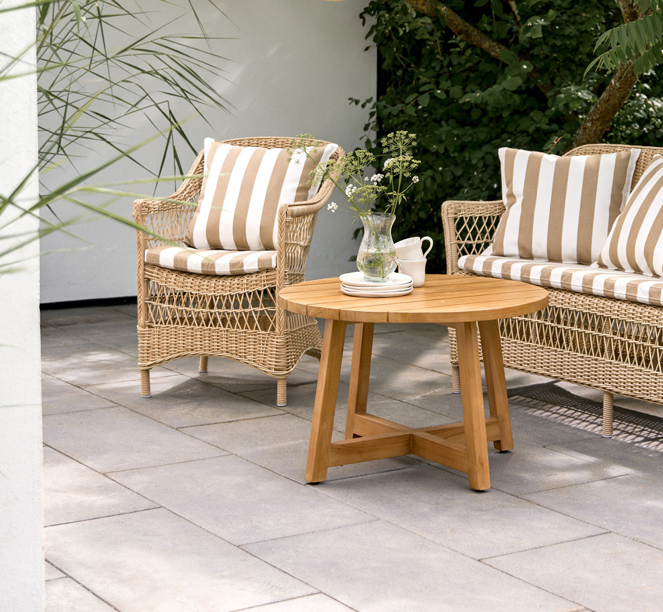 Dies ist ein Lifestyle-Bild von Victor Teak Lounge Table Ø75 cm in teak outdoor. Auf einer Strandterrasse zwischen Tagesbetten mit gestreiften Kissen aufgestellt.