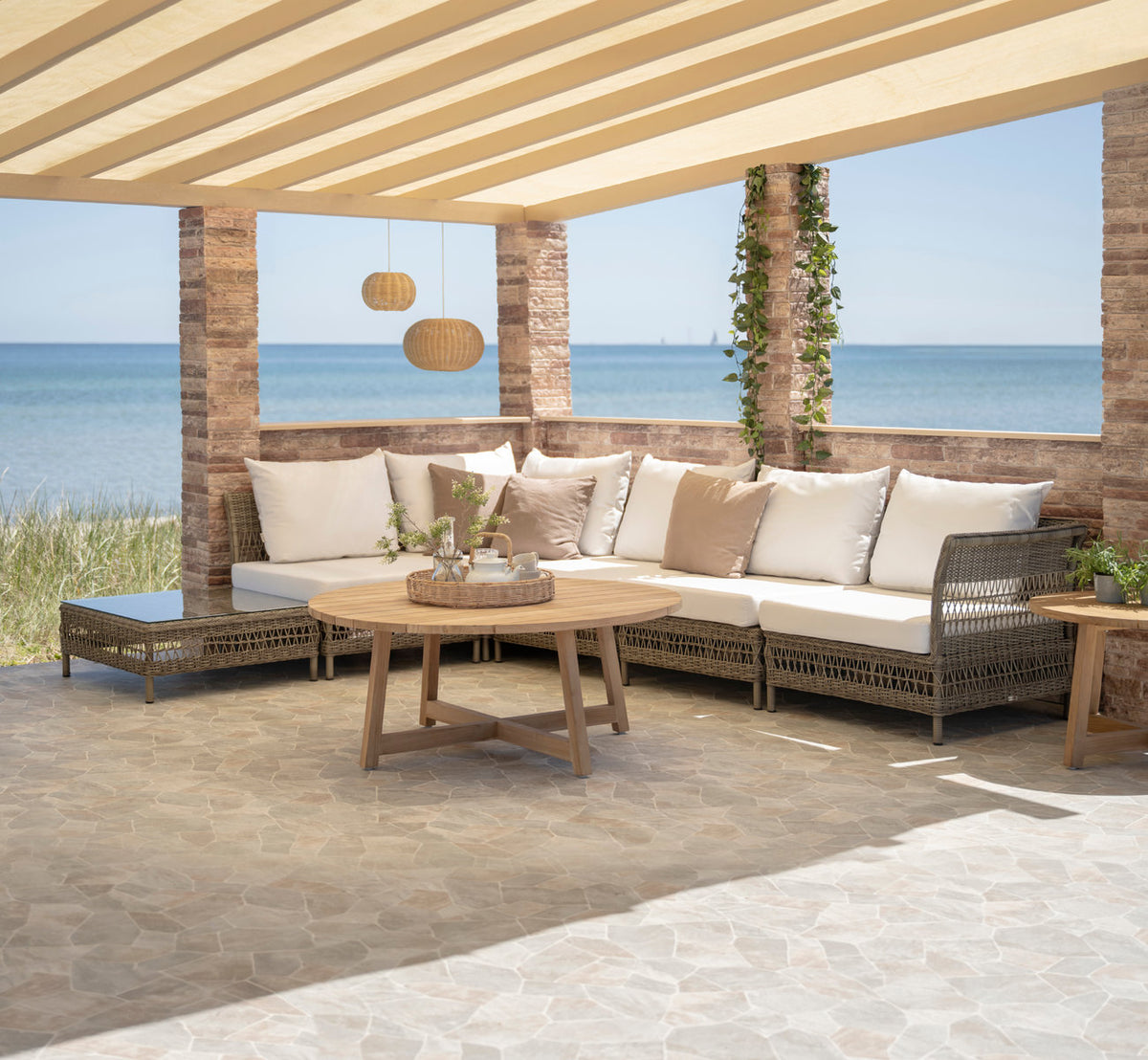 Dies ist ein Lifestyle-Bild von Victor Teak-Loungetisch Ø75 cm in teak outdoor. Ecksofa mit Teaktischen unter einer Pergola am Meer.