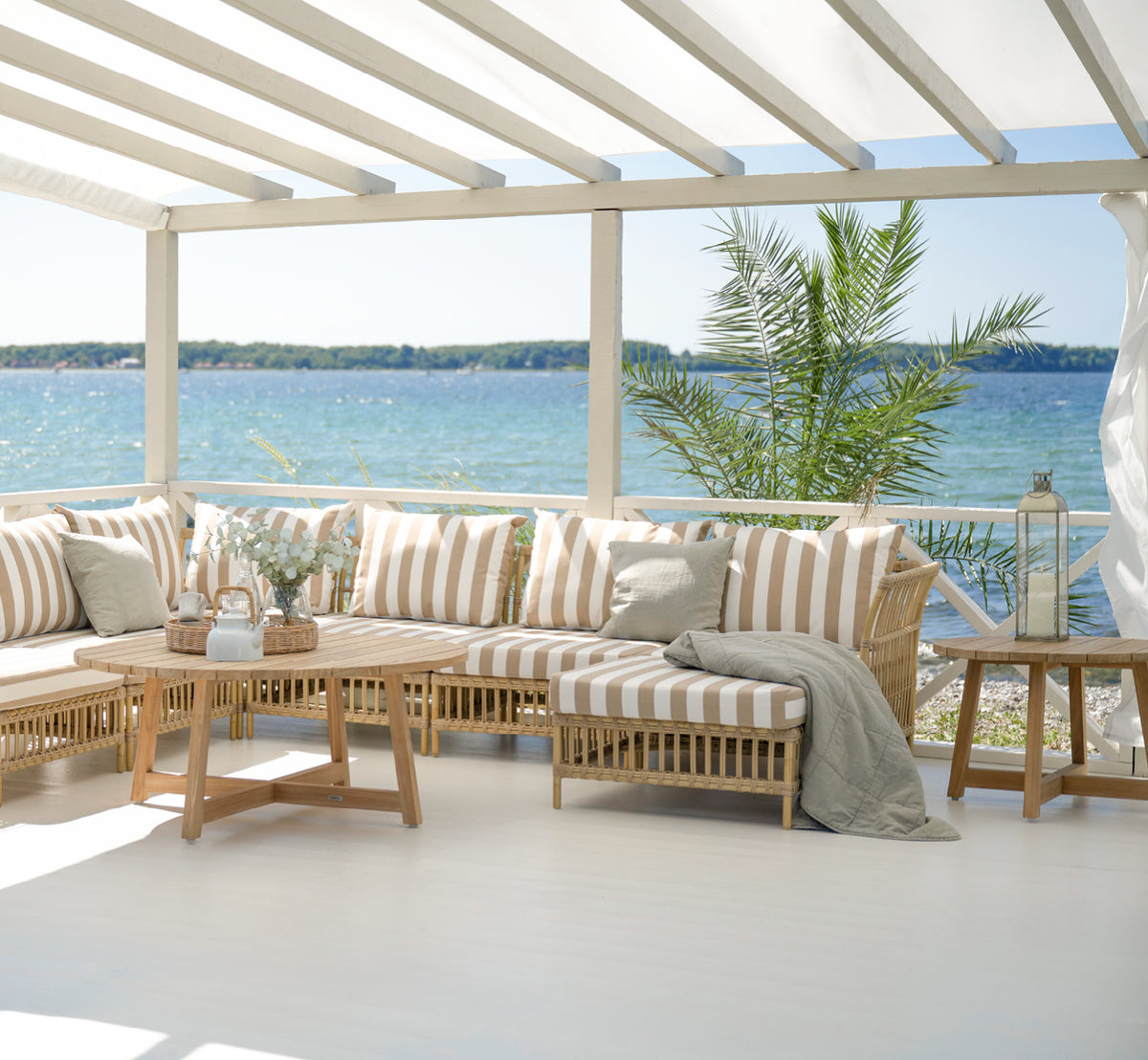 Dies ist ein Lifestyle-Bild von Victor Teak Lounge Table Ø120 cm in teak outdoor. Zwischen Außensofas mit gestreiften Kissen mit Meerblick platziert.