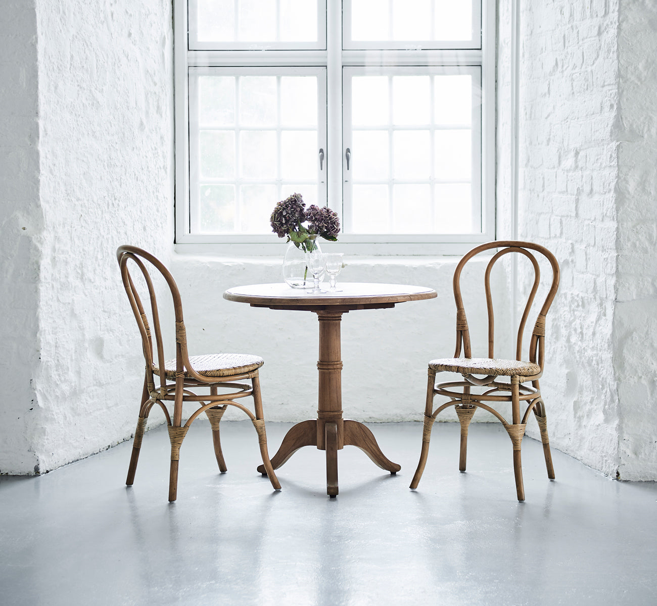 Dies ist ein Lifestyle-Bild von Michel Teak Dining Table Ø80 cm in teak indoor. Mit zwei Stühlen und Blumen in einem hellen weißen Backsteininterieur eingerichtet.