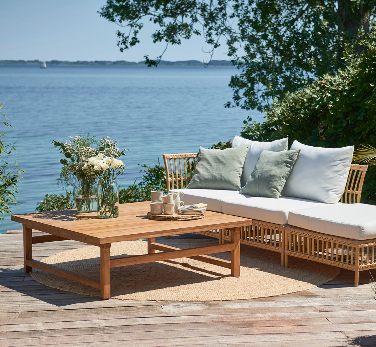Dies ist ein Lifestyle-Bild von Julian Teak Lounge Table 120x120 cm in teak outdoor. Auf einer Strandterrasse mit einem Loungesofa, Kissen und einem Tee-Setting.