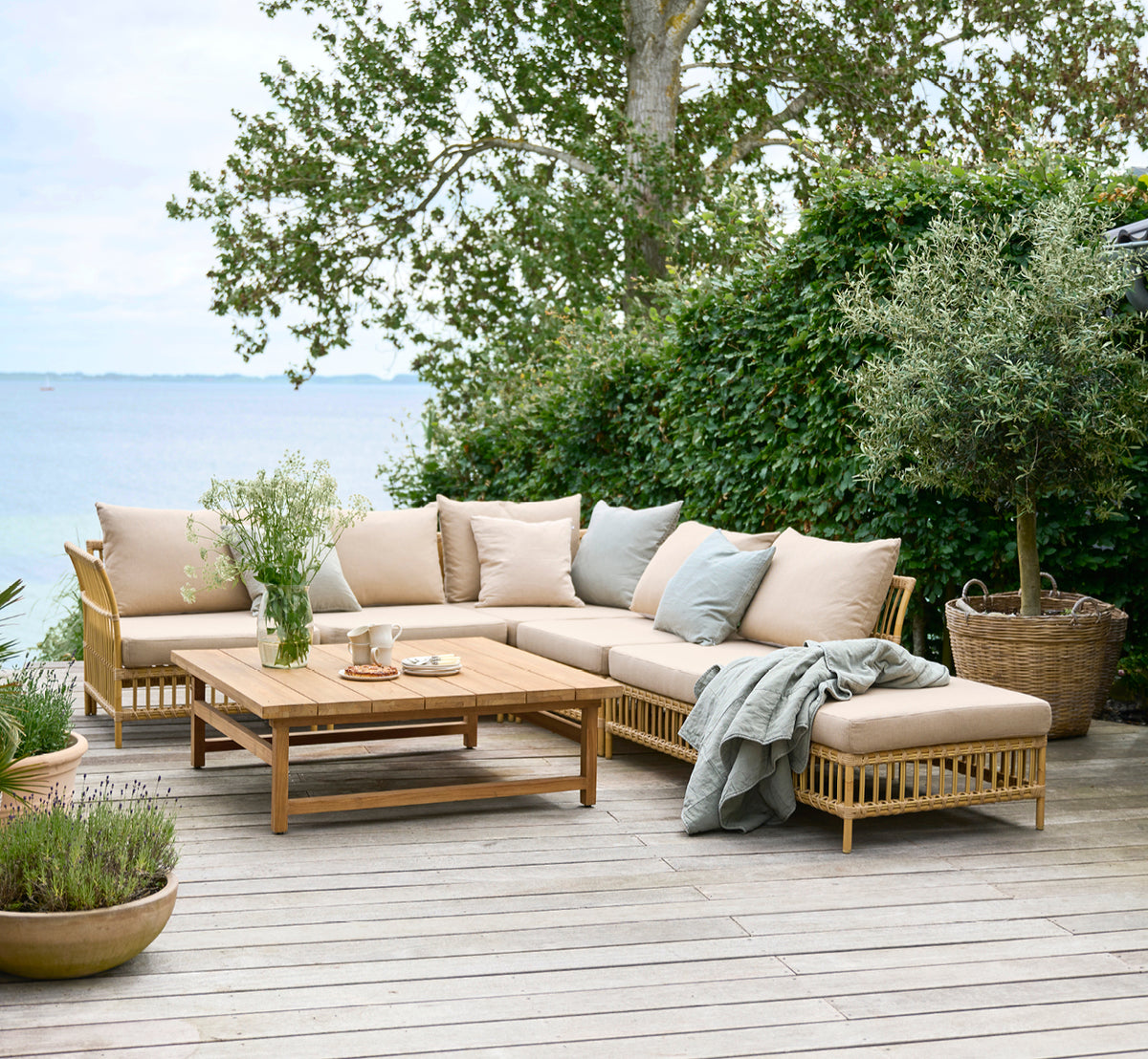 Dies ist ein Lifestyle-Bild von Julian Teak Lounge Table 120x120 cm in teak outdoor. Auf einer Gartenterrasse am Meer mit Loungesitzplätzen und üppigem Grün.