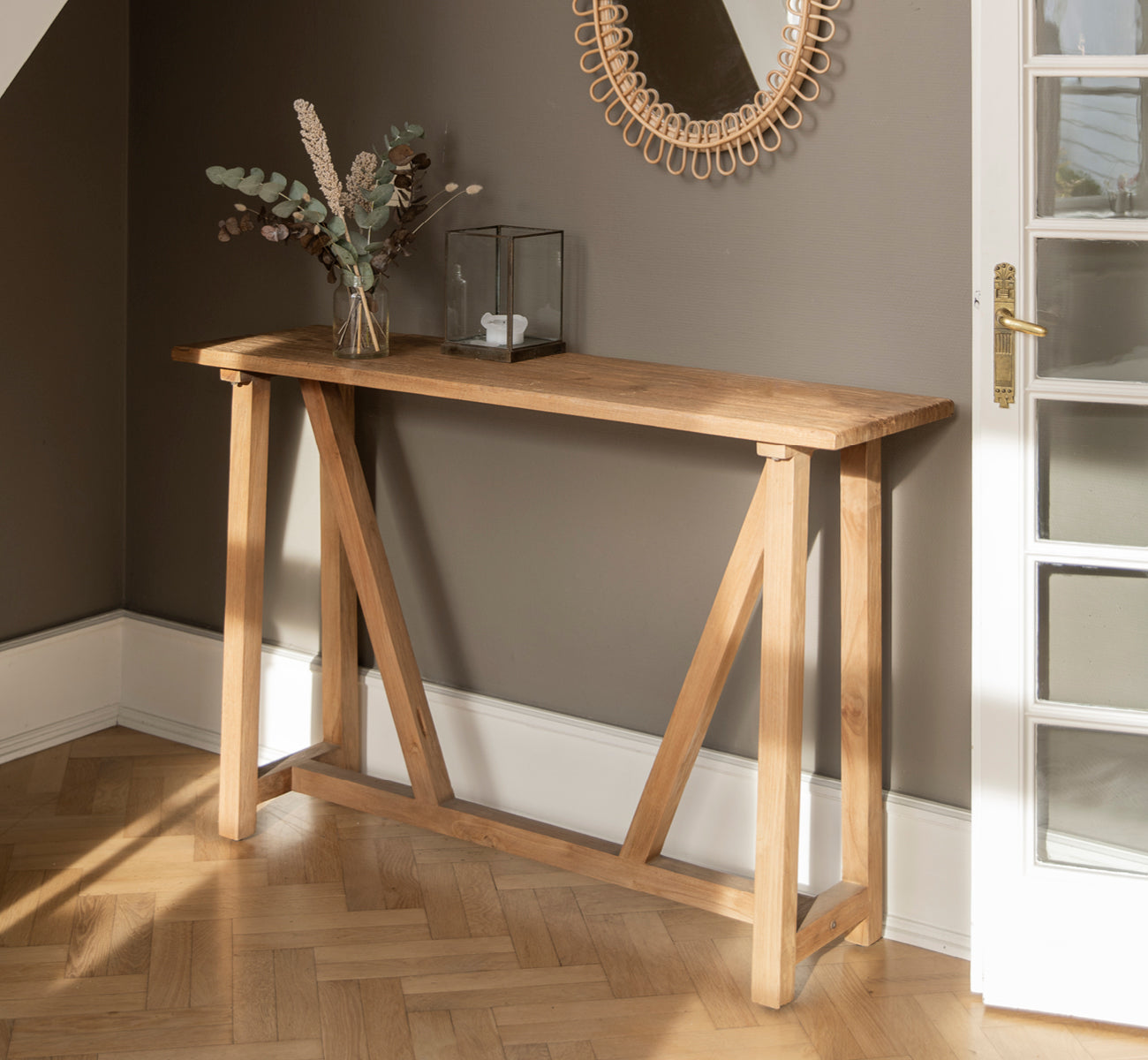 Dies ist ein Lifestyle-Bild von Lucas Teak Konsolentisch in teak indoor. An einer dunkelgrauen Wand mit einem Rattanspiegel darüber und Eukalyptus in einer Vase.
