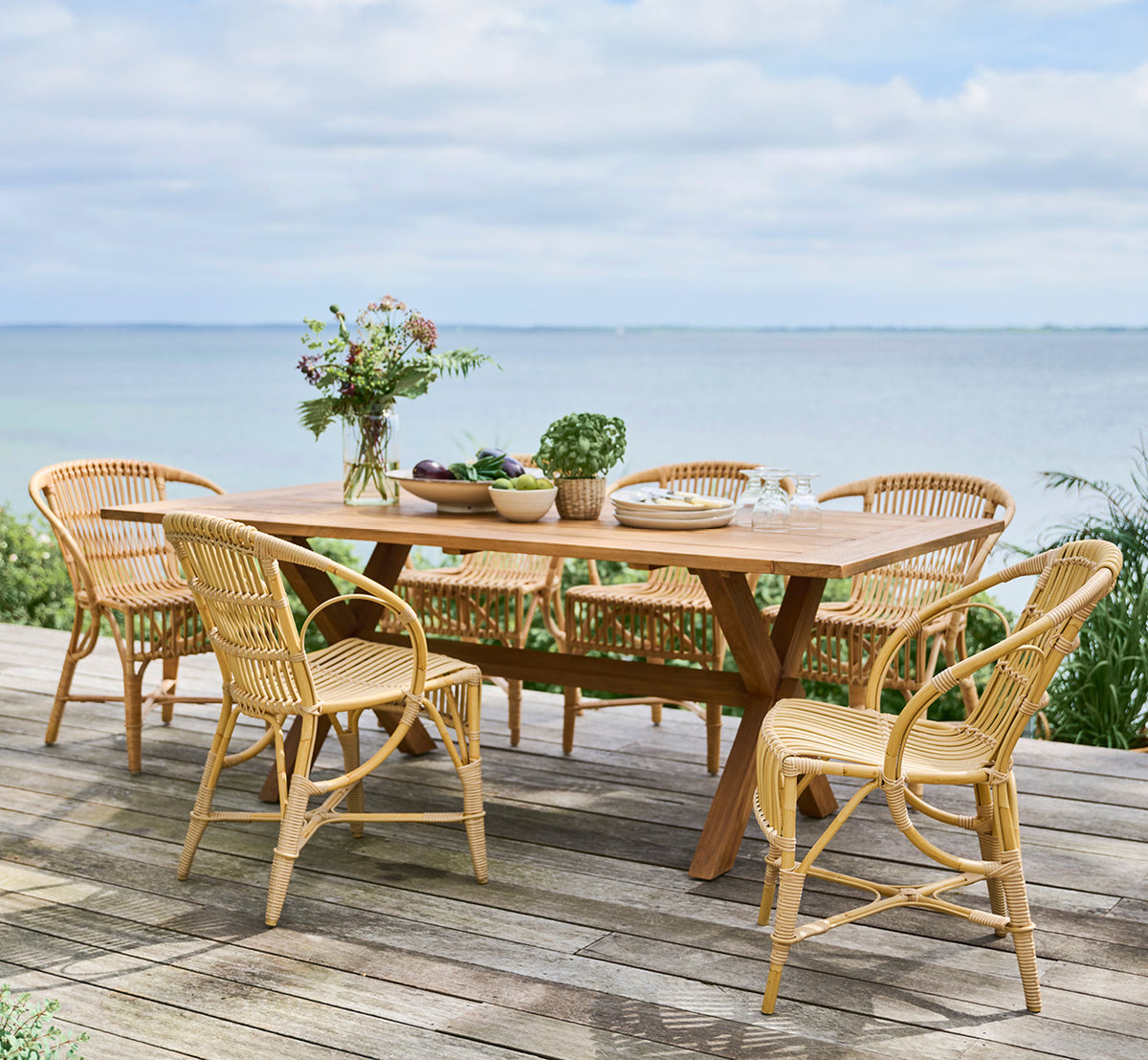 Dies ist ein Lifestyle-Bild von Colonial Tisch 200 in outdoor Teak, auf einer Strandterrasse mit weißem Holzdeck und Meerblick.