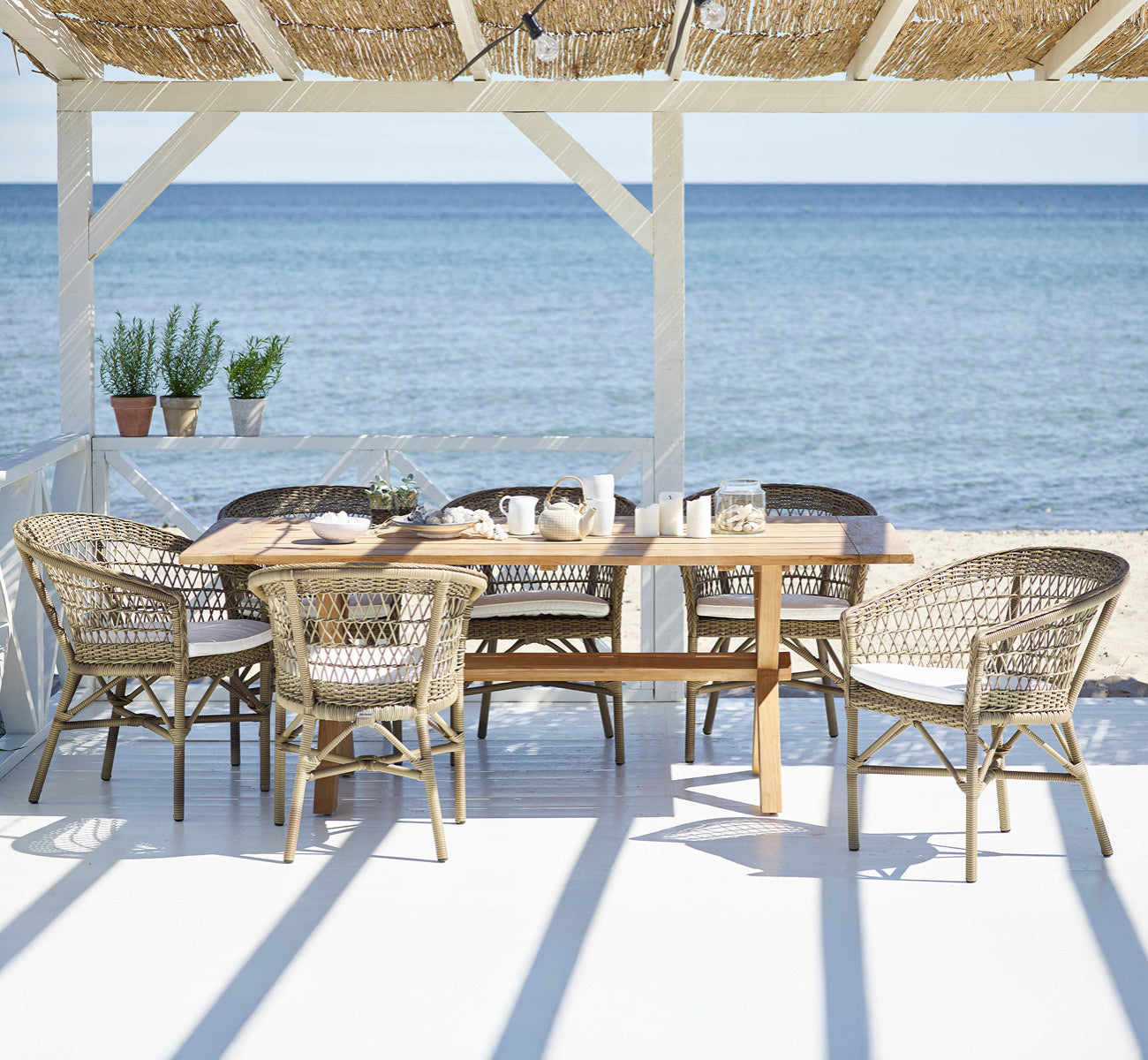 Dies ist ein Lifestyle-Bild von Colonial Tisch 200 in outdoor Teak, auf einer Strandterrasse mit weißem Holzdeck und Meerblick.