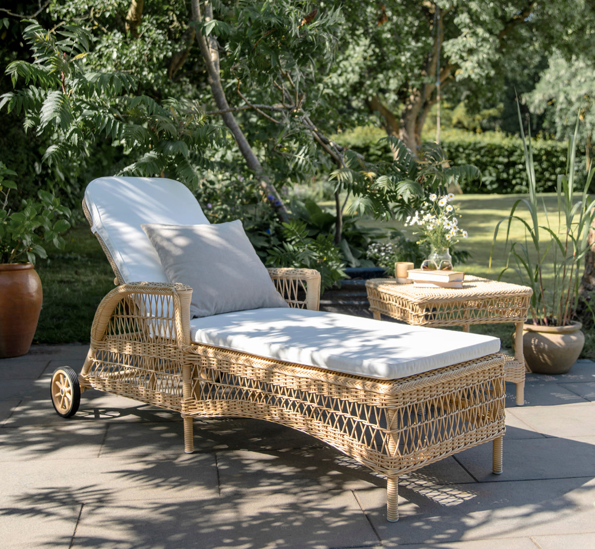 Dies ist ein Lifestyle-Bild von Daisy Sonnenliege in natural ArtFibre Aluminium, auf einer Gartenterrasse umgeben von üppigen Bäumen und Topfpflanzen.