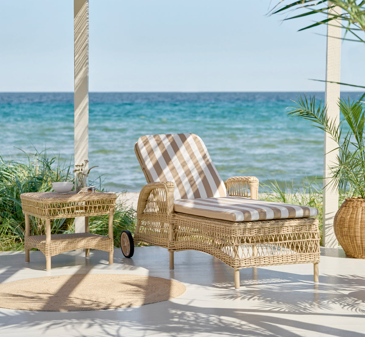 Dies ist ein Lifestyle-Bild von Daisy Sonnenliege in natural ArtFibre Aluminium, auf einer Gartenterrasse umgeben von üppigen Bäumen und Topfpflanzen.