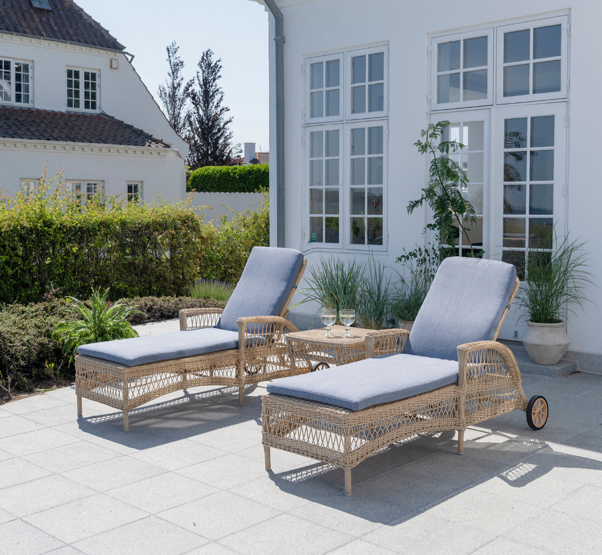 Dies ist ein Lifestyle-Bild von Daisy Sonnenliege in natural ArtFibre Aluminium, auf einer Gartenterrasse umgeben von üppigen Bäumen und Topfpflanzen.