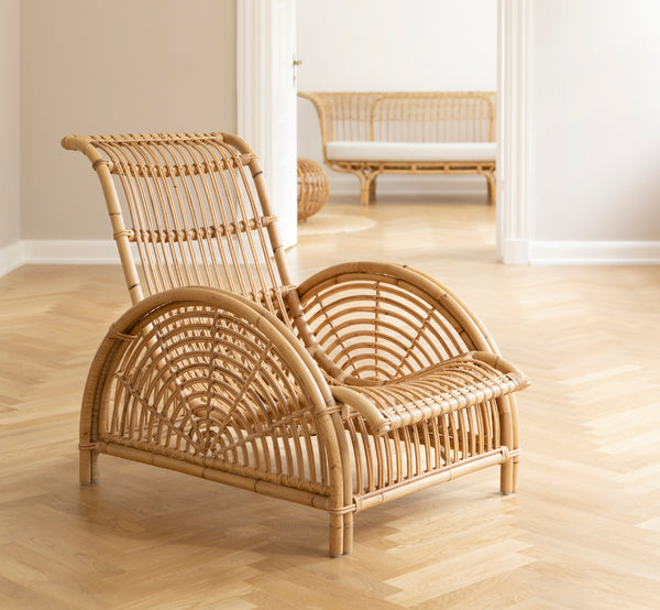Dies ist ein Lifestyle-Bild von Paris Lounge-Sessel in polished natural rattan. Auf einem Fischgrätparkettboden neben einer passenden Bank platziert.