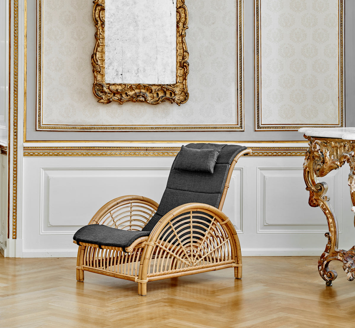 Dies ist ein Lifestyle-Bild von Paris Lounge-Sessel in polished natural rattan. Auf einem Fischgrätparkettboden neben einer passenden Bank platziert.