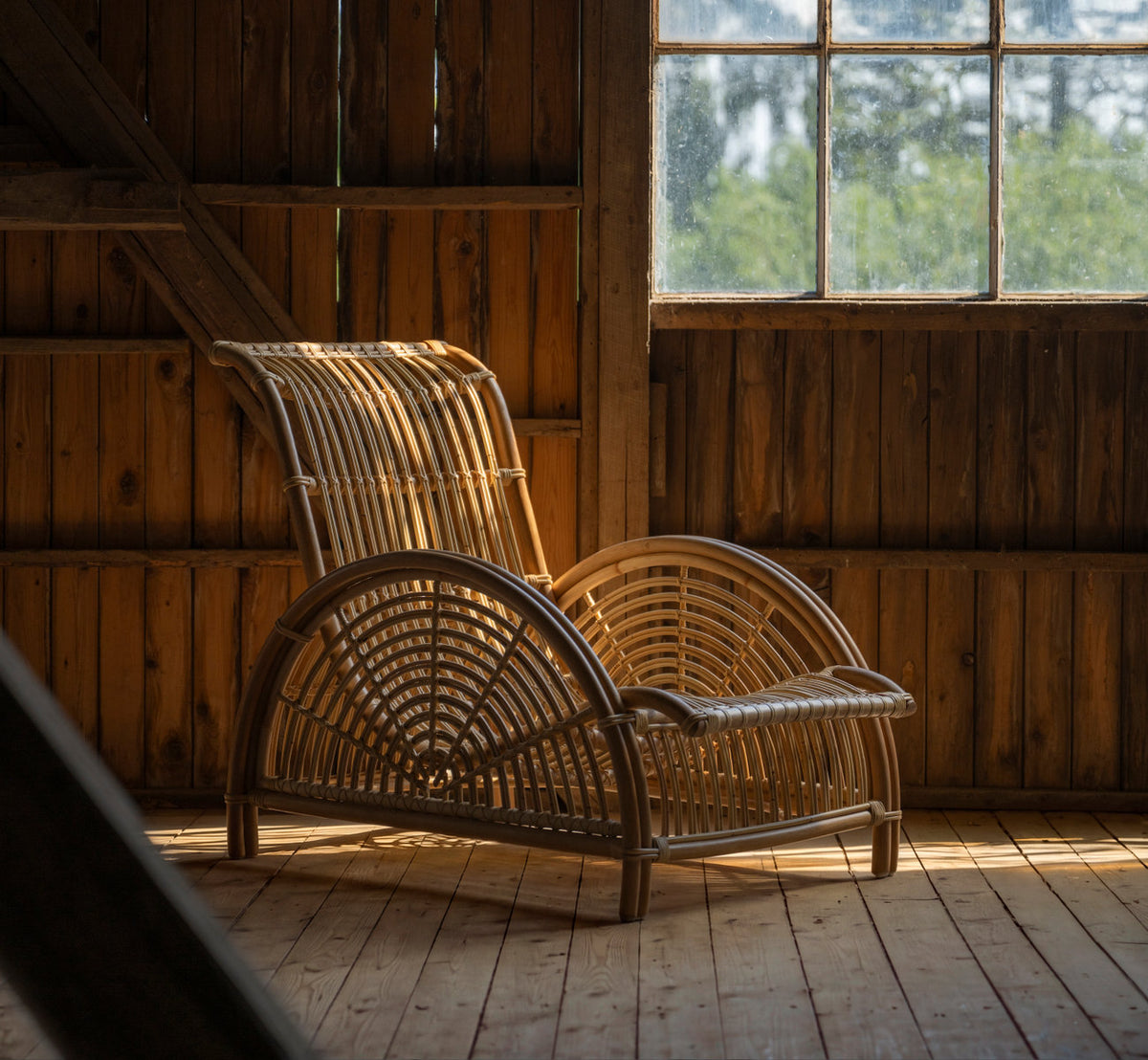 Dies ist ein Lifestyle-Bild von Paris Lounge-Sessel in polished natural rattan. In einer Holzscheune im warmen Nachmittagslicht.