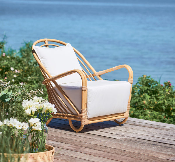 Dies ist ein Lifestyle-Bild von Charlottenborg Außen-Lounge-Sessel in natural Aluminium ArtFibre, auf einer Holzterrasse im Freien mit Blumen und.