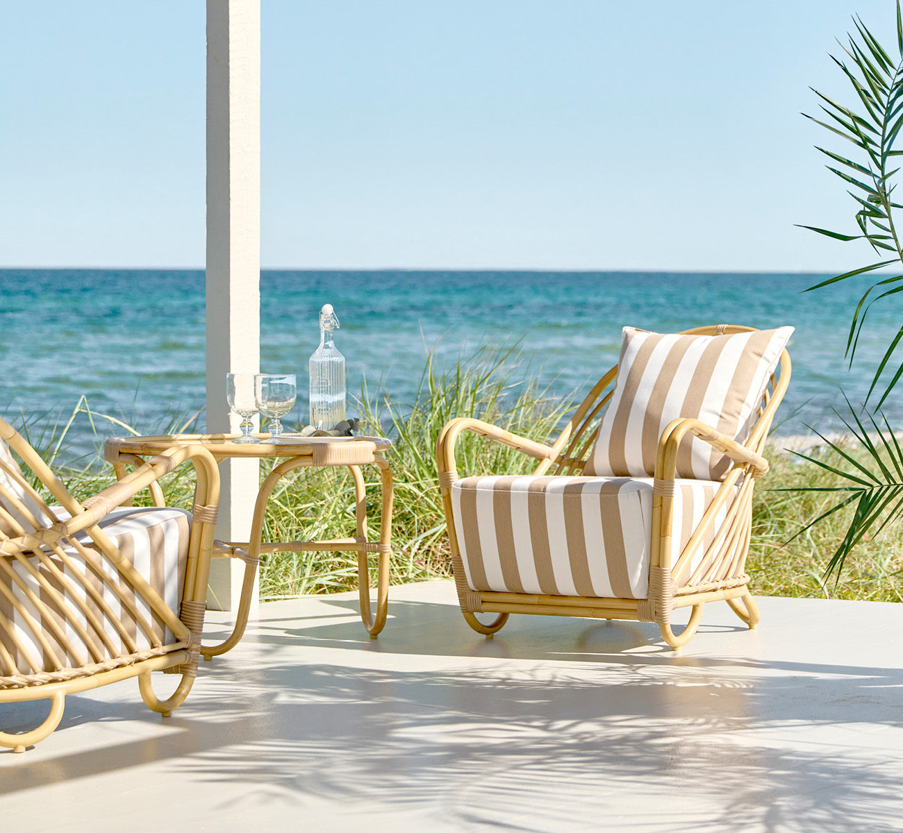 Dies ist ein Lifestyle-Bild von Charlottenborg Außen-Lounge-Sessel in natural Aluminium ArtFibre, auf einer Holzterrasse im Freien mit Blumen und.