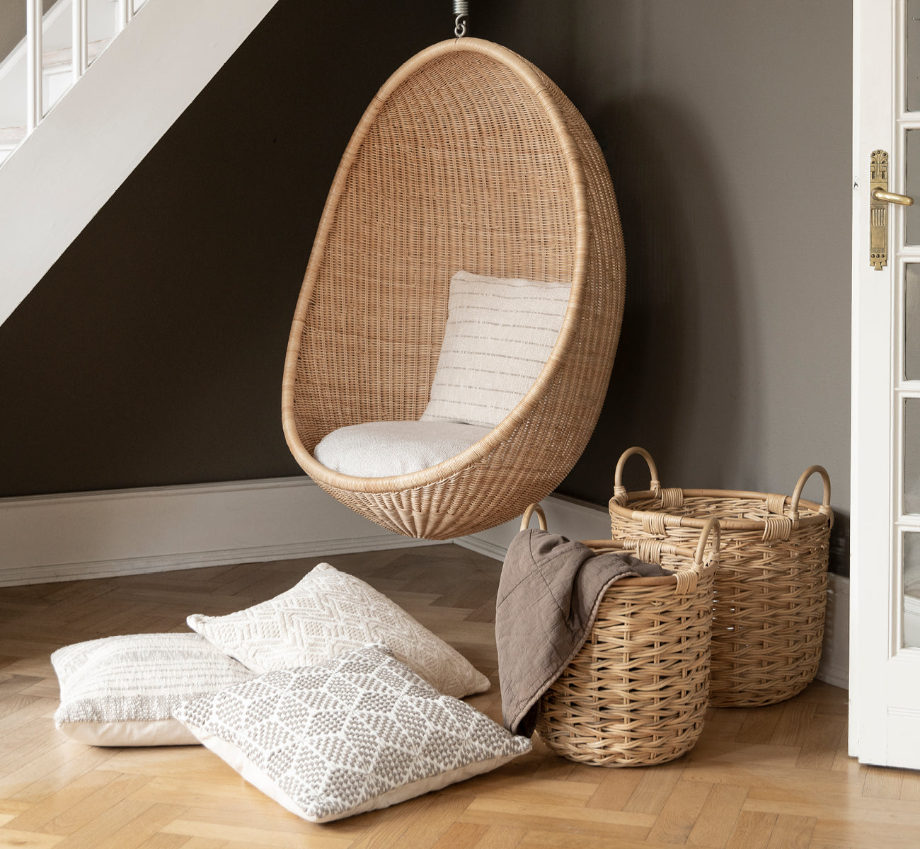 Dies ist ein Lifestyle-Bild von Sarah Korb-Set in natural rattan. Neben einem hängenden Hängesessel und gemusterten Kissen auf Parkett platziert.