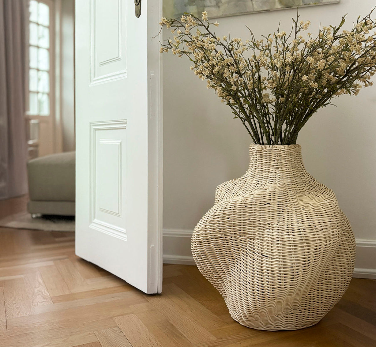 Dies ist ein Lifestyle-Bild von Solaris Basket Ø46 cm in natural core weave iron frame. Mit Trockenblumen neben einer weißen Tür auf Parkett gestylt.