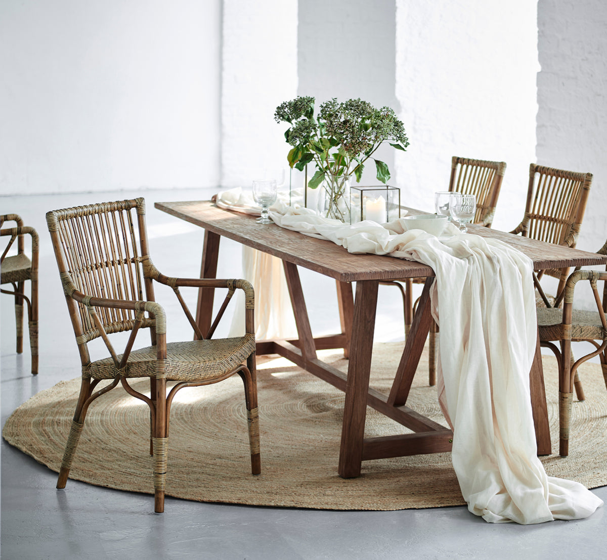 Dies ist ein Lifestyle-Bild von Jute Carpet Ø280 in grey jute. Unter einem Esstisch mit Rattanstühlen, Leinenstoff und Grün.