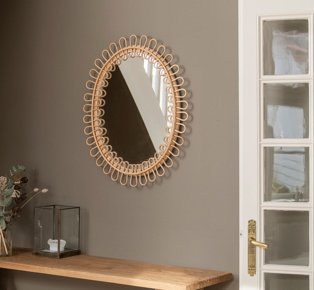 Dies ist ein Lifestyle-Bild von Luella Spiegel in polished natural rattan. An einer grauen Wand über einem Holzregal mit Eukalyptus und Glasdekoration hängend.