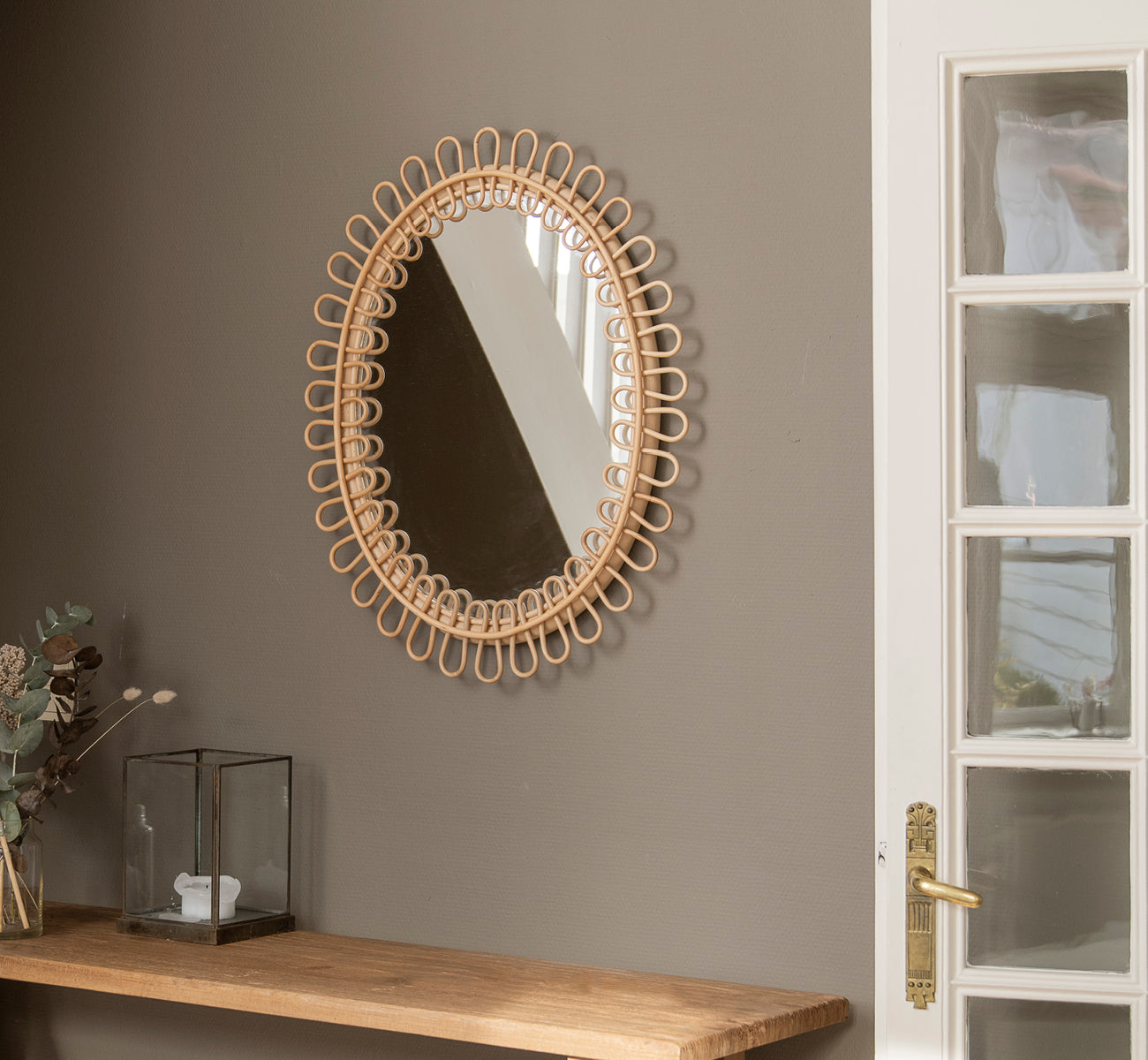 Dies ist ein Lifestyle-Bild von Luella Spiegel in polished natural rattan. An einer grauen Wand über einem Holzregal mit Eukalyptus und Glasdekoration hängend.