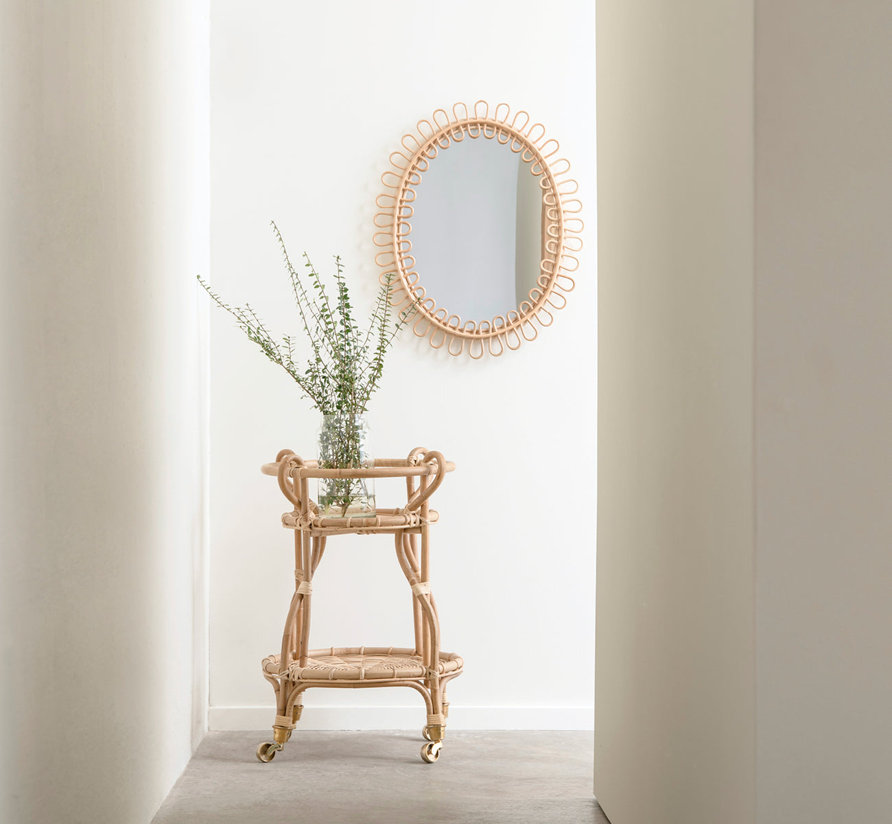 Dies ist ein Lifestyle-Bild von Luella Spiegel in polished natural rattan. In einer hellen Flurnische über einem Rattan-Barwagen mit frischem Grün.
