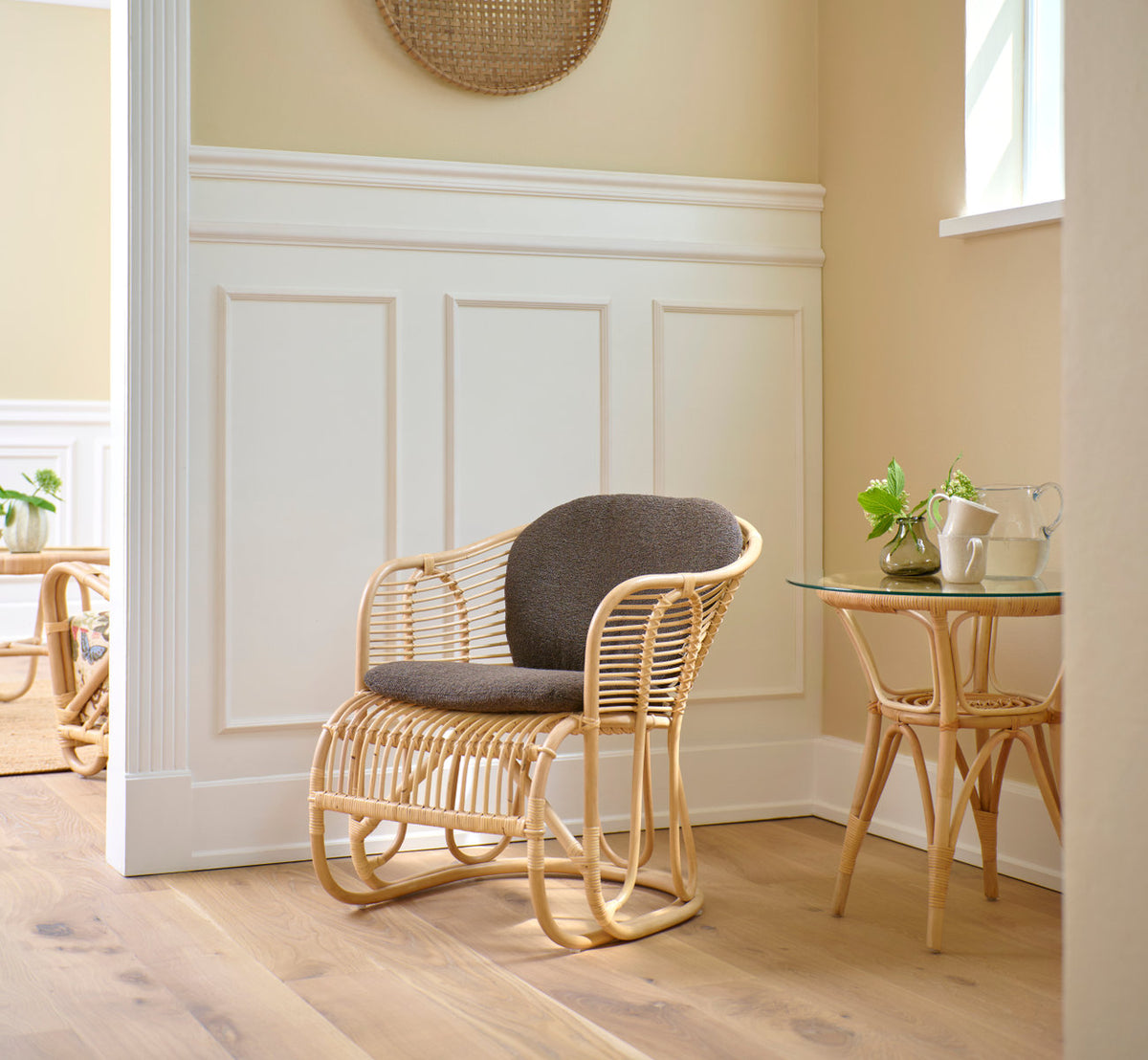 Dies ist ein Lifestyle-Bild von Swing Lounge-Sessel in polished natural rattan. Mit grauem Kissen in einer hellen, vertäfelten Ecke.