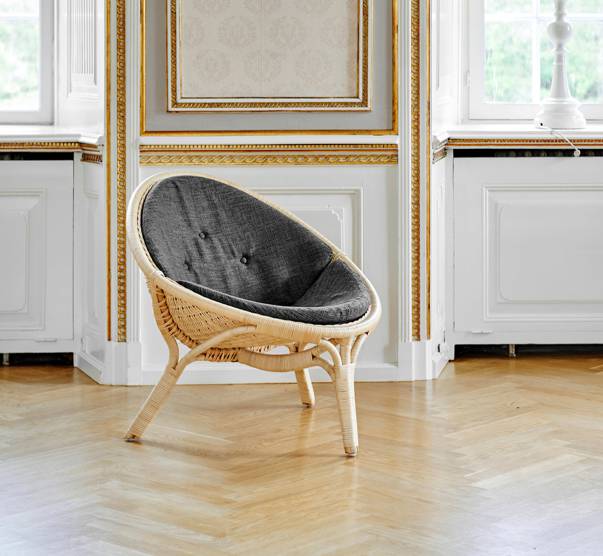 Dies ist ein Lifestyle-Bild von Rana Lounge-Sessel in natural core weave rattan frame. Allein in einem Raum mit vergoldeter Vertäfelung und Parkettboden stehend.