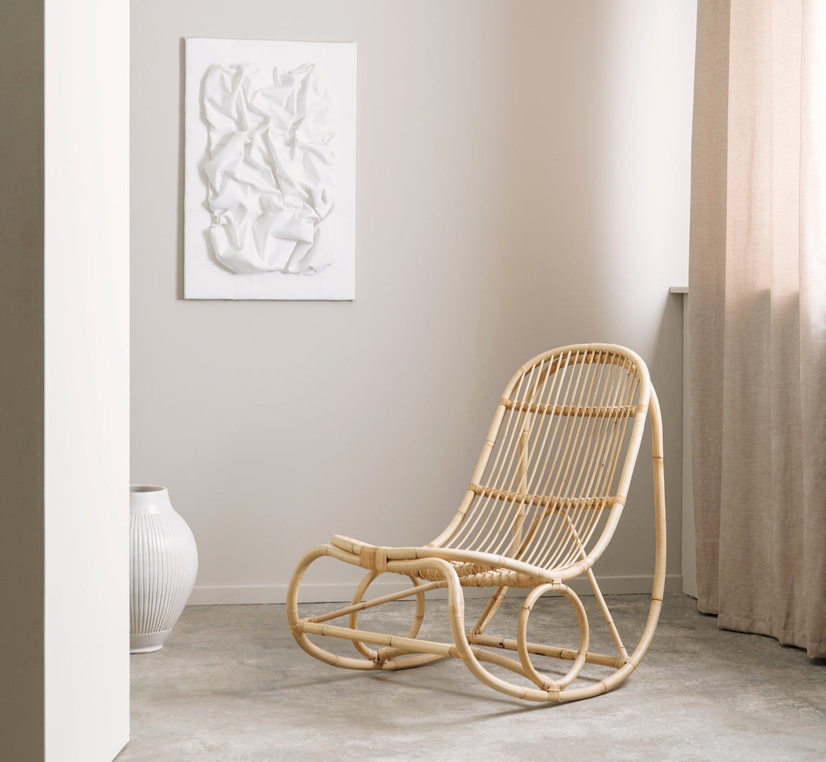 Dies ist ein Lifestyle-Bild von Nanny Rocking Chair i polished natural rattan. In einer Galerie-Umgebung mit anderen Rattanmöbeln auf Sockeln.