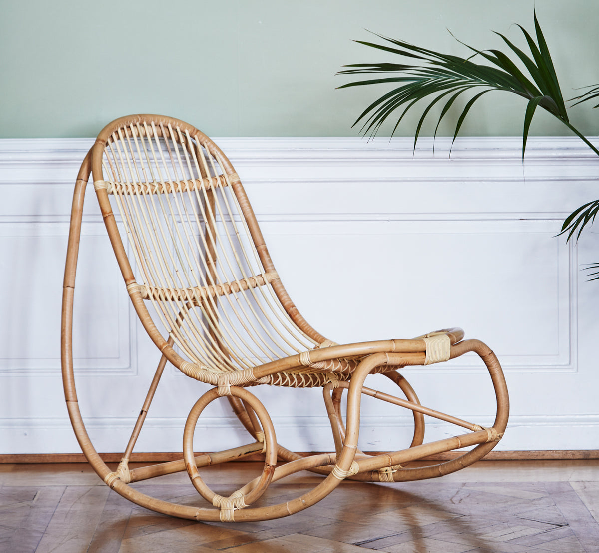 Dies ist ein Lifestyle-Bild von Nanny Rocking Chair i polished natural rattan. Vor einer salbeigrünen Wand mit weißer Vertäfelung und Palme.