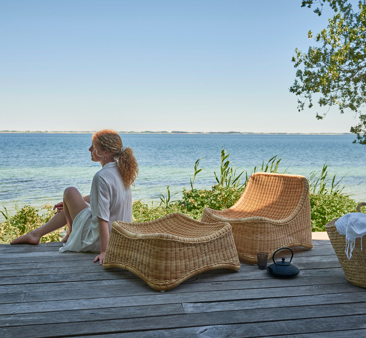 Dies ist ein Lifestyle-Bild von Chill Lounge-Sessel in natural core weave ArtFibre Aluminium, auf einem Holzdeck am Wasser mit Küstengrün und.