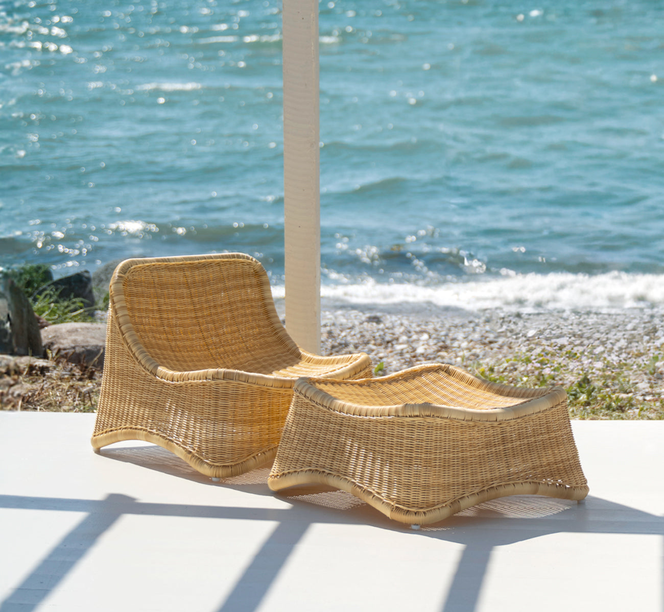 Dies ist ein Lifestyle-Bild von Chill Lounge-Sessel in natural core weave ArtFibre Aluminium, auf einem Holzdeck am Wasser mit Küstengrün und.
