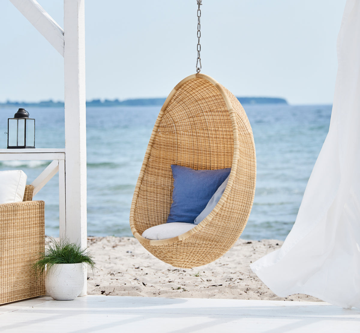 Dies ist ein Lifestyle-Bild von Hanging Außen-Hängesessel in natural core weave ArtFibre Aluminium, hängend an einer weißen Pergola mit Sandstrand.