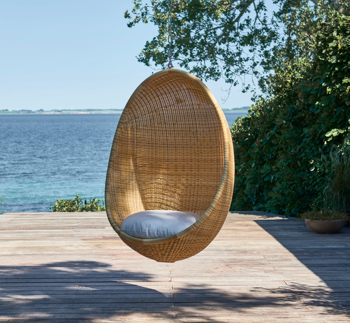 Dies ist ein Lifestyle-Bild von Hanging Außen-Hängesessel in natural core weave ArtFibre Aluminium, hängend an einer weißen Pergola mit Sandstrand.