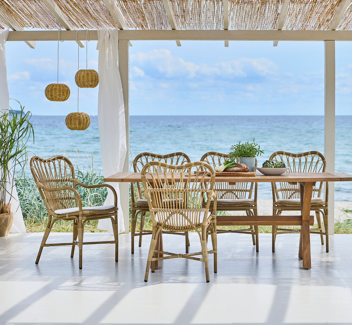 Dies ist ein Lifestyle-Bild von Margret Außen-Esszimmer-Armlehnstuhl in antique aluminium artfibre. Strandpavillon mit Pendelleuchten und Meerblick.