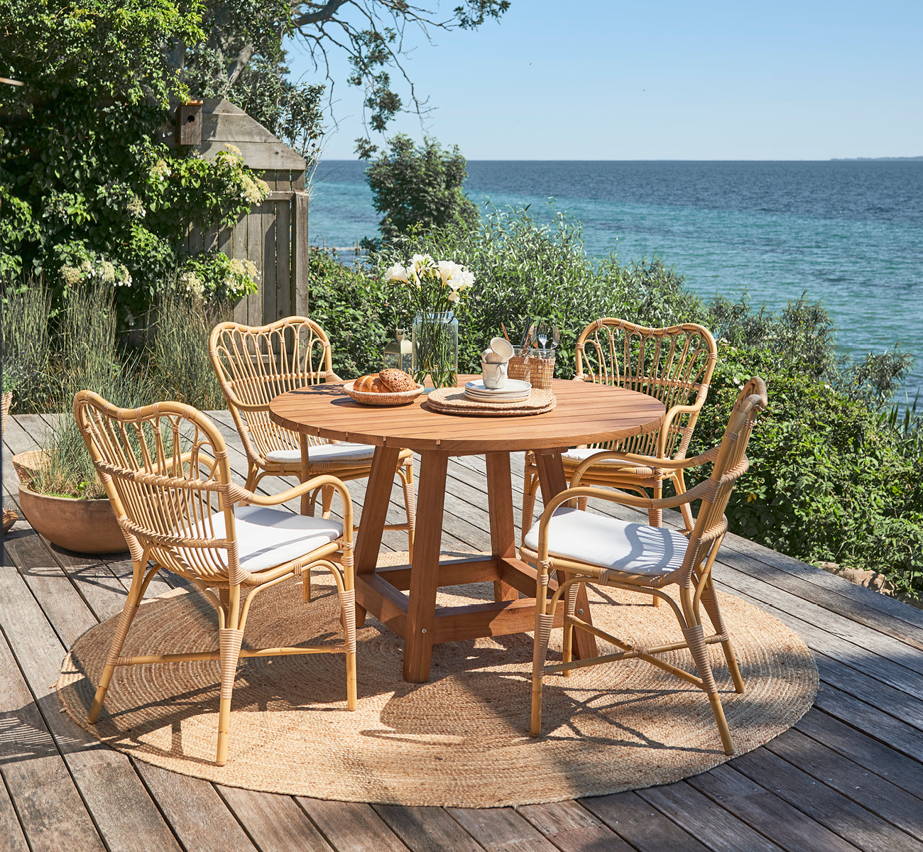 Dies ist ein Lifestyle-Bild von Margret Außen-Esszimmer-Armlehnstuhl in natural aluminium artfibre. Terrasse am Meer mit rundem Teaktisch und Gartenpflanzen.