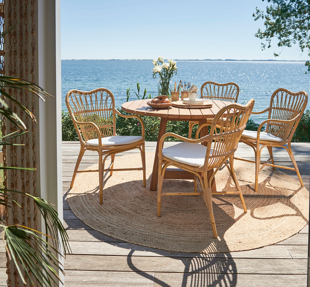 Dies ist ein Lifestyle-Bild von Margret Außen-Esszimmer-Armlehnstuhl in natural aluminium artfibre. Terrasse am Meer mit rundem Teaktisch und Meerblick.