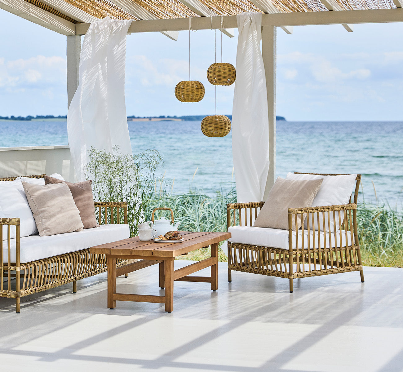 Dies ist ein Lifestyle-Bild von Caroline Außen-Lounge-Sessel in antique Aluminium ArtFibre, auf einem Holzdeck am Wasser mit Küstengrün und Meerblick.