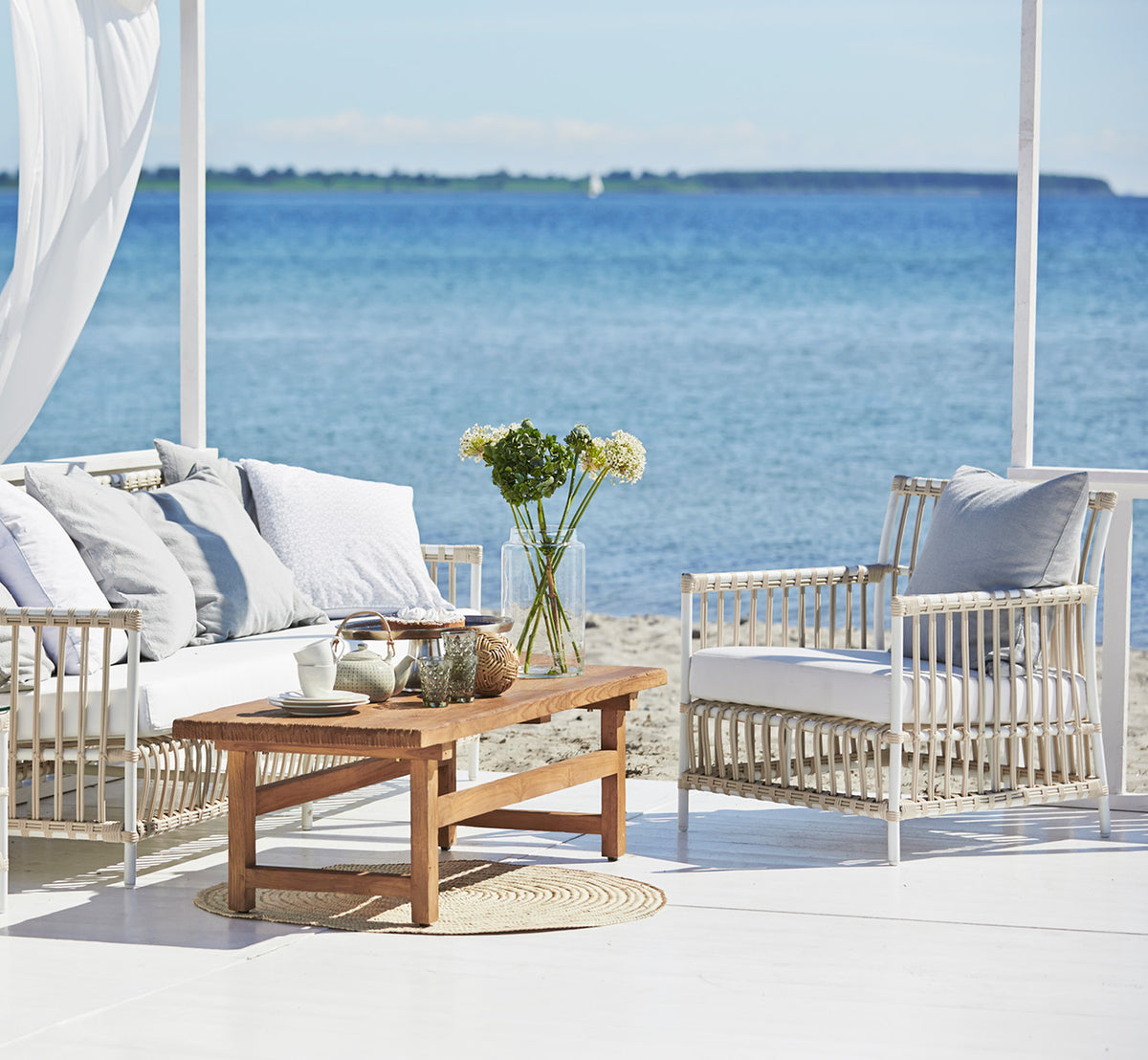 Dies ist ein Lifestyle-Bild von Caroline Außen-Lounge-Sessel in dove White Aluminium ArtFibre, auf einem Holzdeck am Wasser mit Küstengrün und.