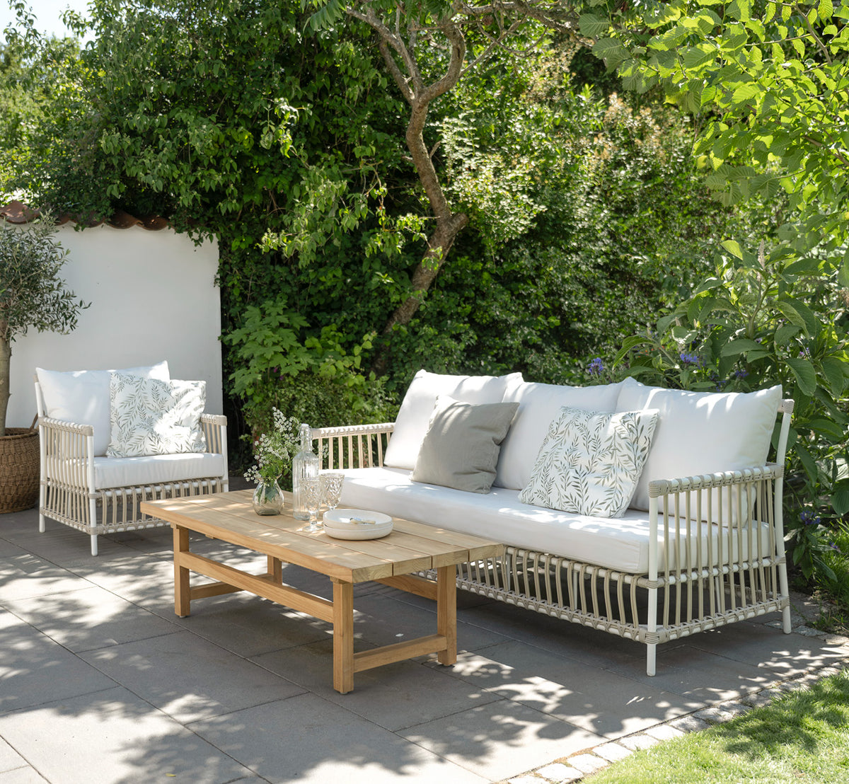 Dies ist ein Lifestyle-Bild von Caroline Außen-Lounge-Sessel in dove White Aluminium ArtFibre, auf einem Holzdeck am Wasser mit Küstengrün und.