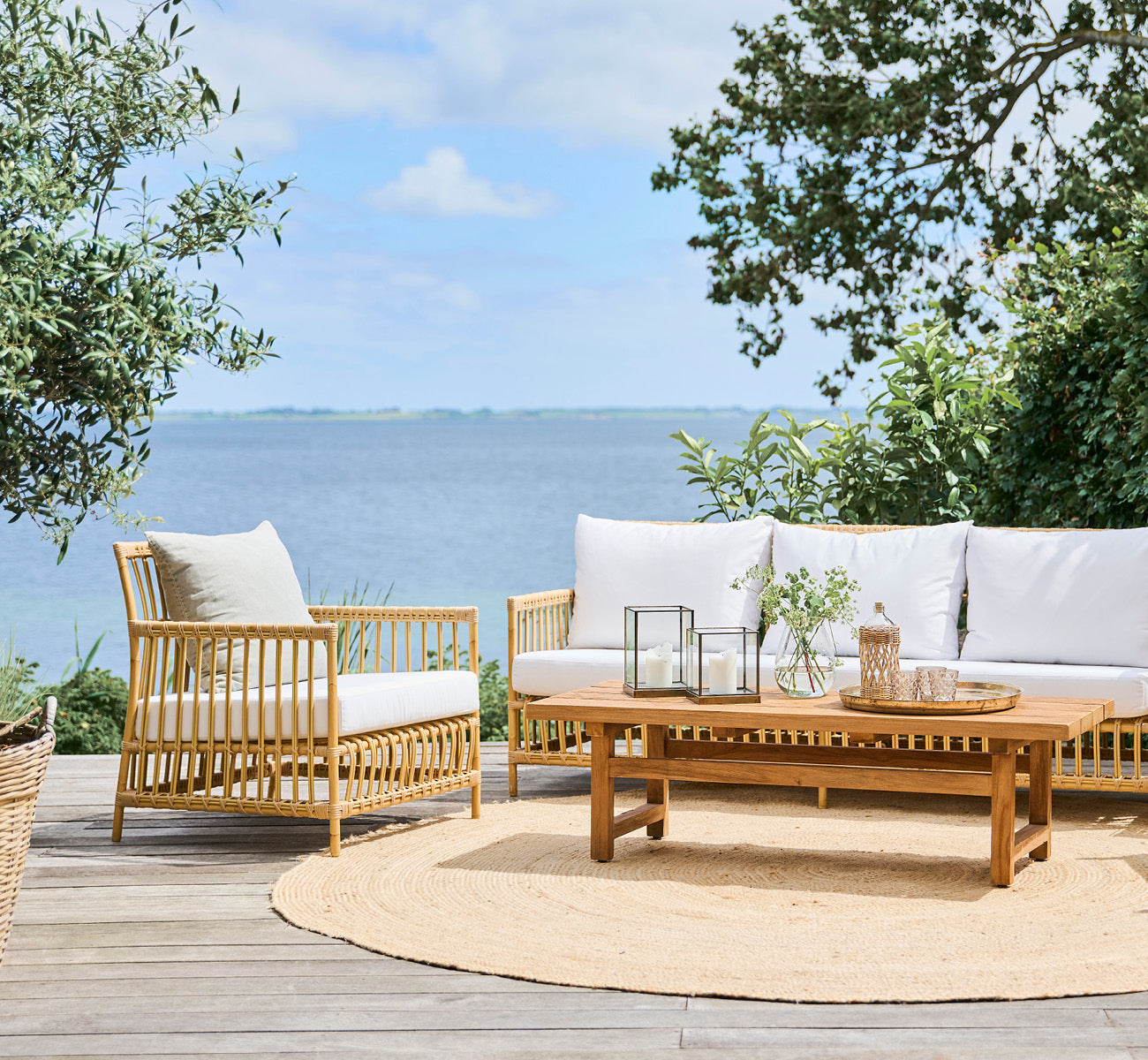 Dies ist ein Lifestyle-Bild von Caroline Außen-Lounge-Sessel in natural Aluminium ArtFibre, auf einem Holzdeck am Wasser mit Küstengrün und Meerblick.