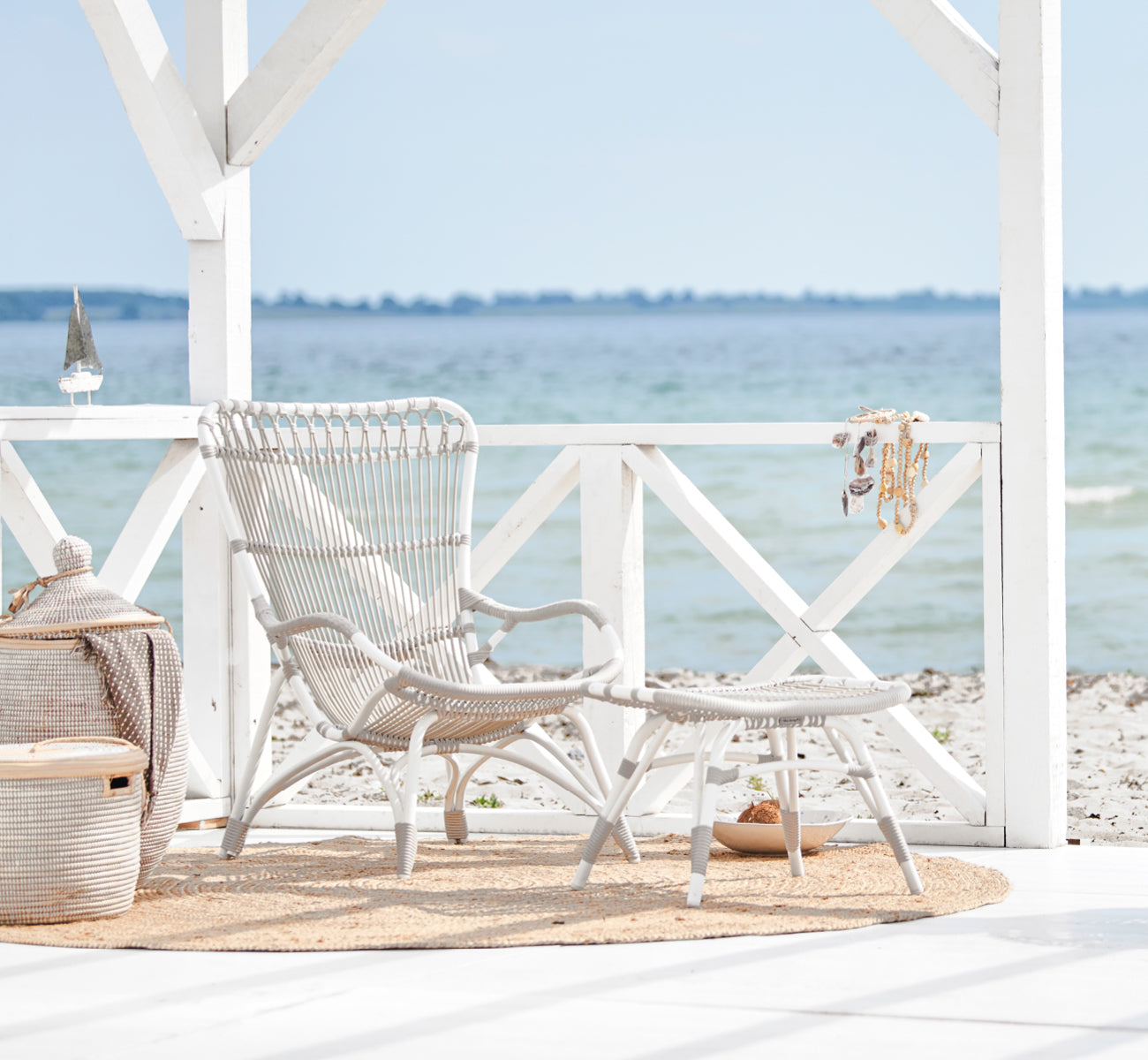 Dies ist ein Lifestyle-Bild von Monet Exterior Footstool i dove white aluminium artfibre. In einem Strandpavillon mit Juteteppich und Korb.