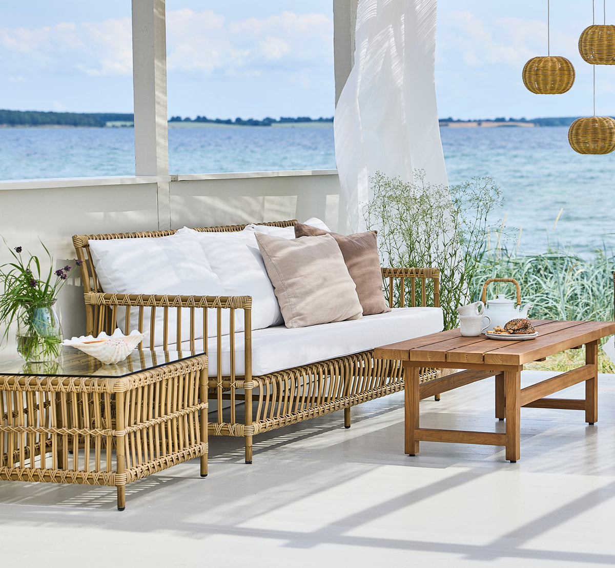 Dies ist ein Lifestyle-Bild von Caroline Außen-Sofa in antique Aluminium ArtFibre, auf einem Holzdeck am Wasser mit Küstengrün und Meerblick.