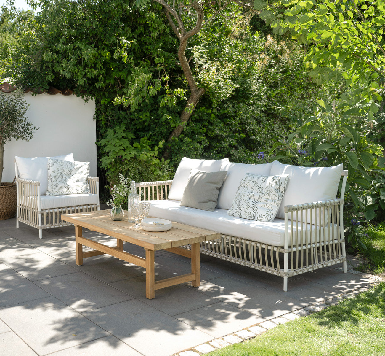Dies ist ein Lifestyle-Bild von Caroline Außen-Sofa in dove White Aluminium ArtFibre, auf einem Holzdeck am Wasser mit Küstengrün und Meerblick.
