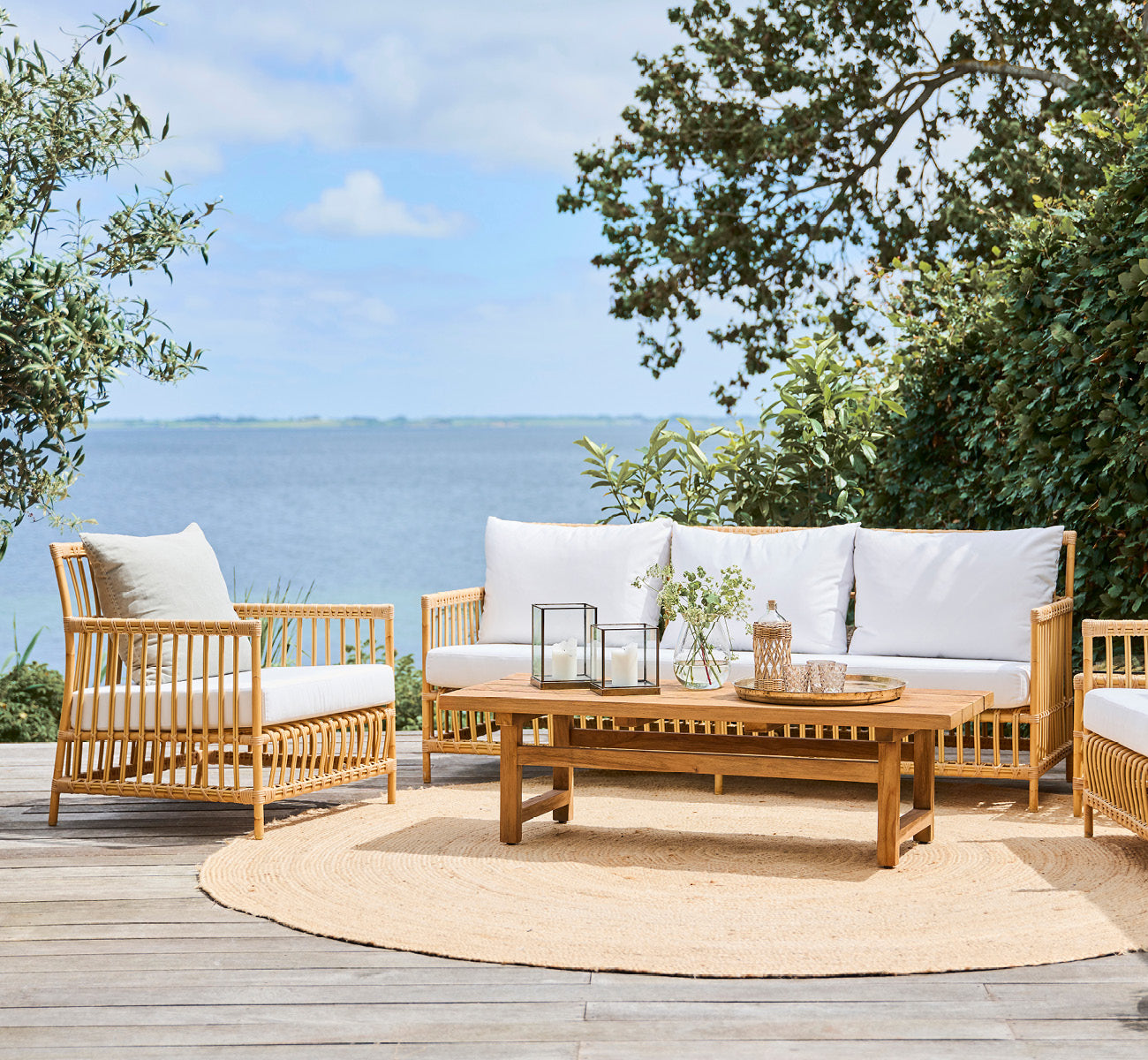Dies ist ein Lifestyle-Bild von Caroline Außen-Sofa in natural Aluminium ArtFibre, auf einem Holzdeck am Wasser mit Küstengrün und Meerblick.