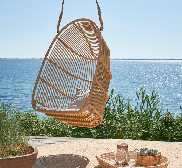 Dies ist ein Lifestyle-Bild von Renoir Außen-Hängesessel in natural aluminium artfibre. Auf einer Strandterrasse mit Tablett und Sukkulenten darunter hängend.