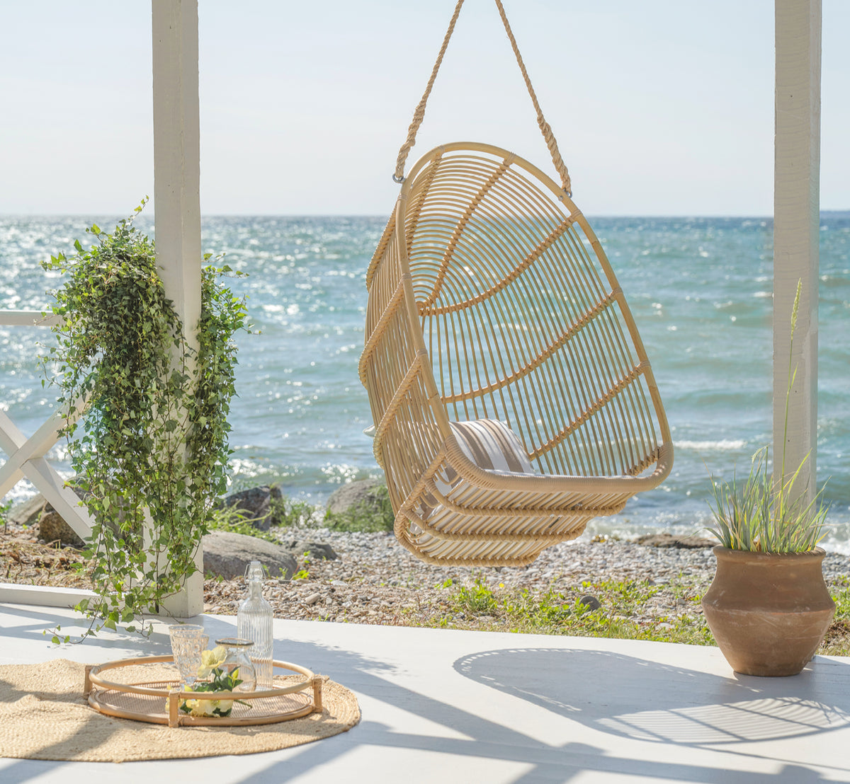 Dies ist ein Lifestyle-Bild von Renoir Außen-Hängesessel in natural aluminium artfibre. Auf einer Strandterrasse mit Tablett und Sukkulenten darunter hängend.